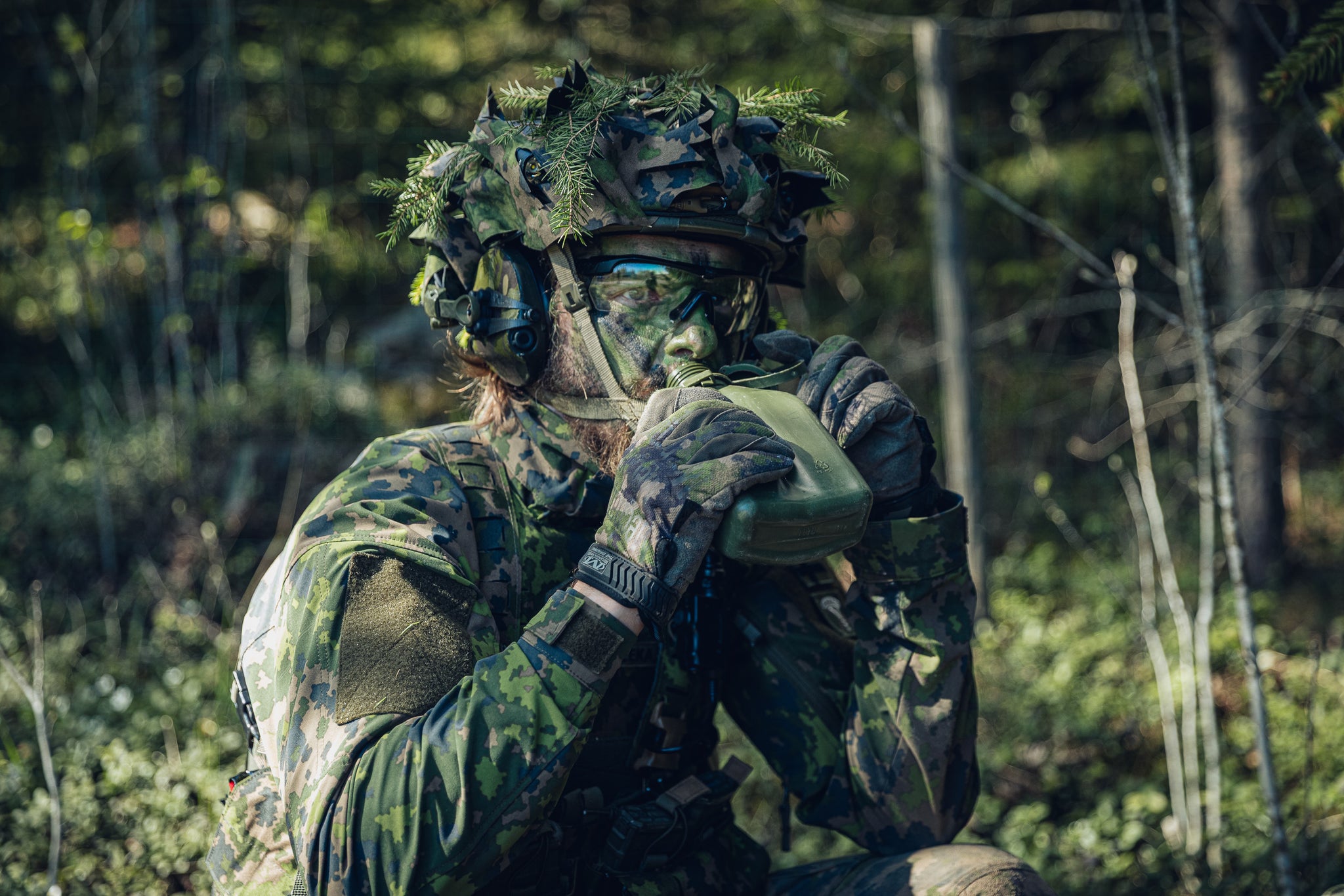 A person in full Finnish M05 camouflage military-style gear, including a ghillie helmet, face paint, and gloves, drinking from a green canteen in a forest setting.