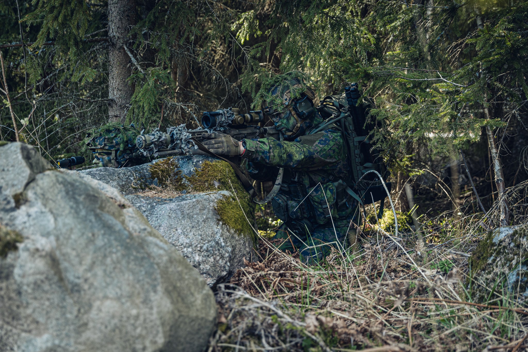 A person in full camouflage tactical gear crouches behind rocks in a dense forest, aiming a rifle. 