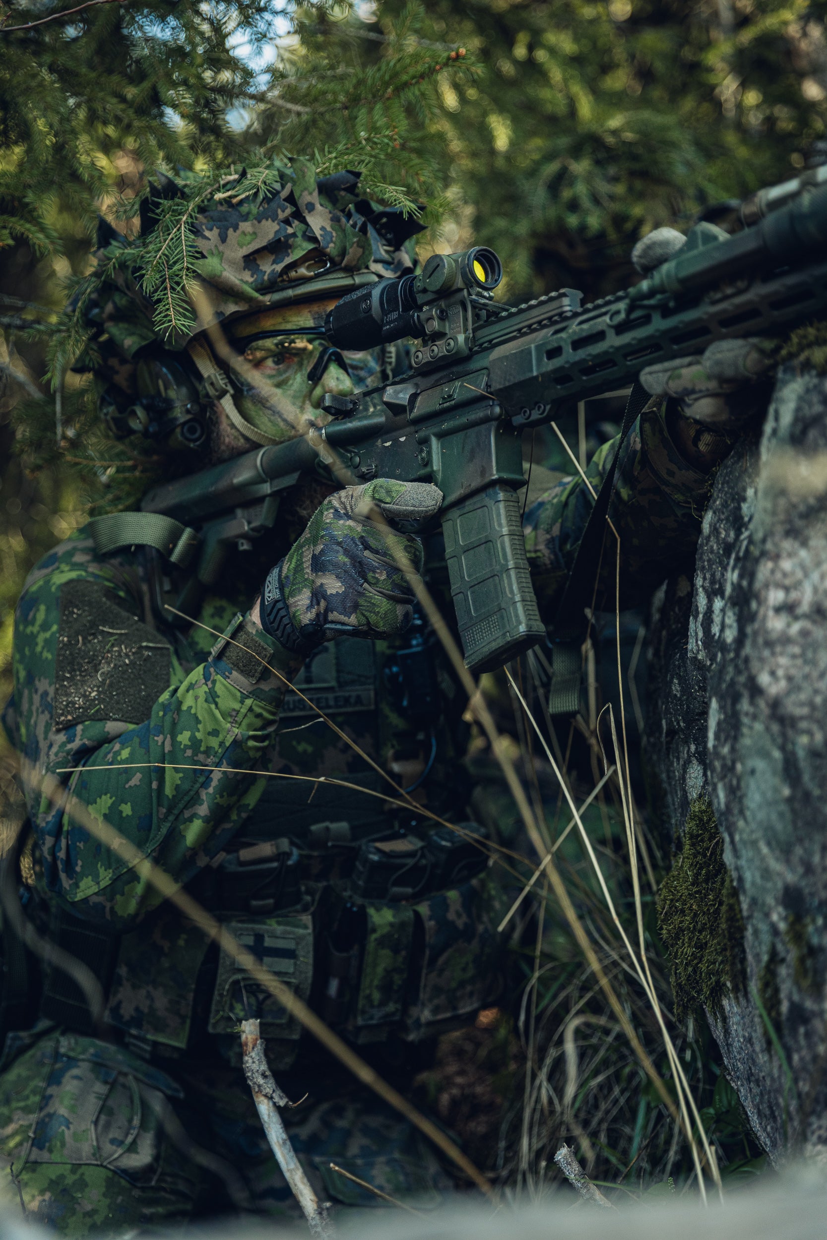 A person in full camouflage tactical gear and a leafy ghillie head covering aims a black rifle with a mounted scope in a dense forest setting.