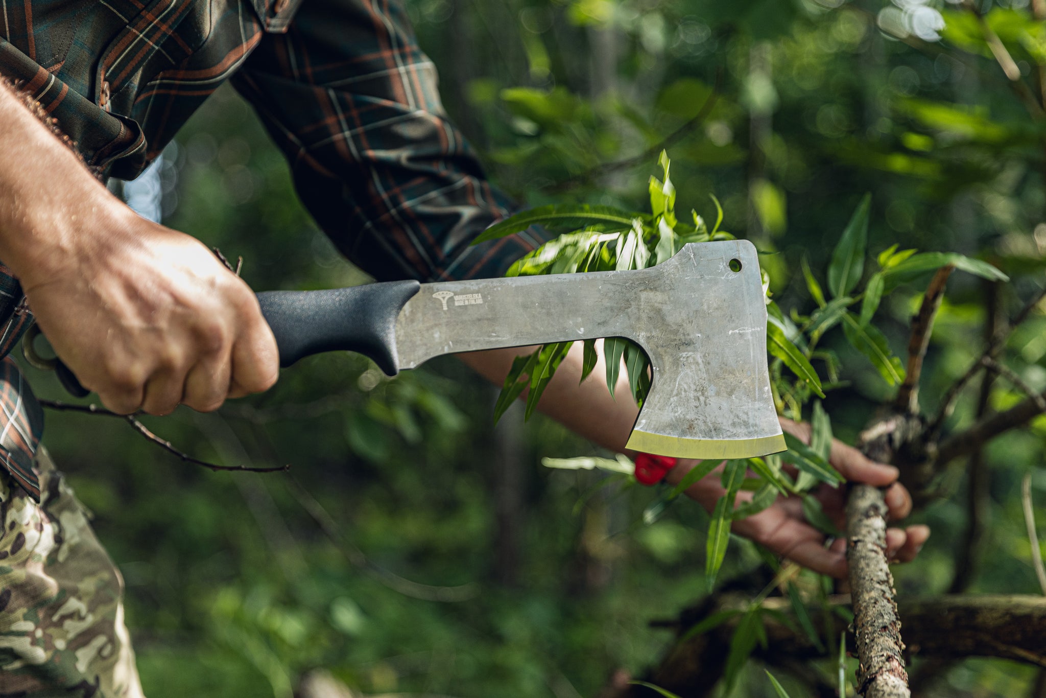 A close-up of a person holding a black-handled axe in their right hand and a stick in the other in a forest setting.