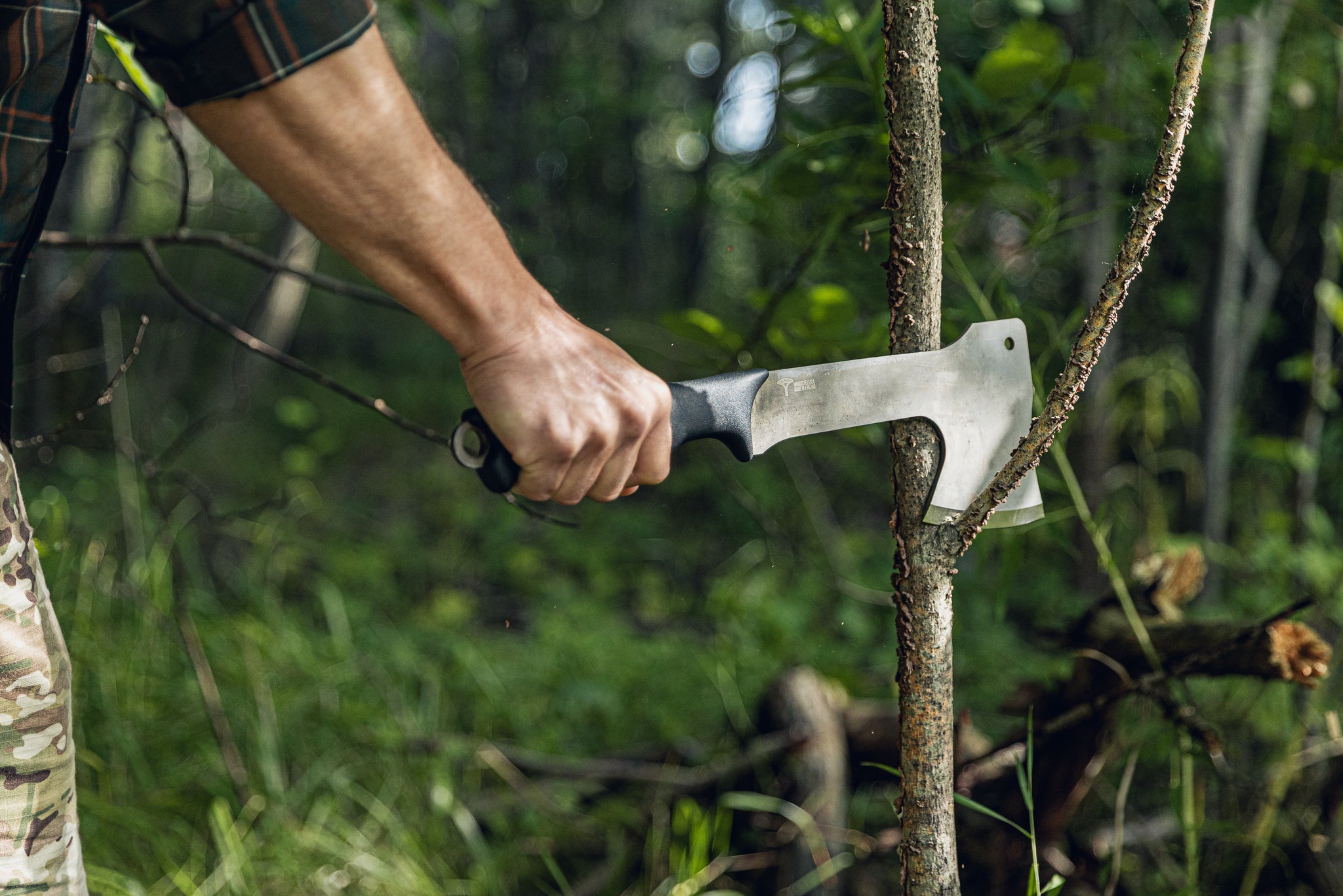 A close-up action shot of a person's hand holding a hatchet and splitting a tree trunk in a dense green forest.
