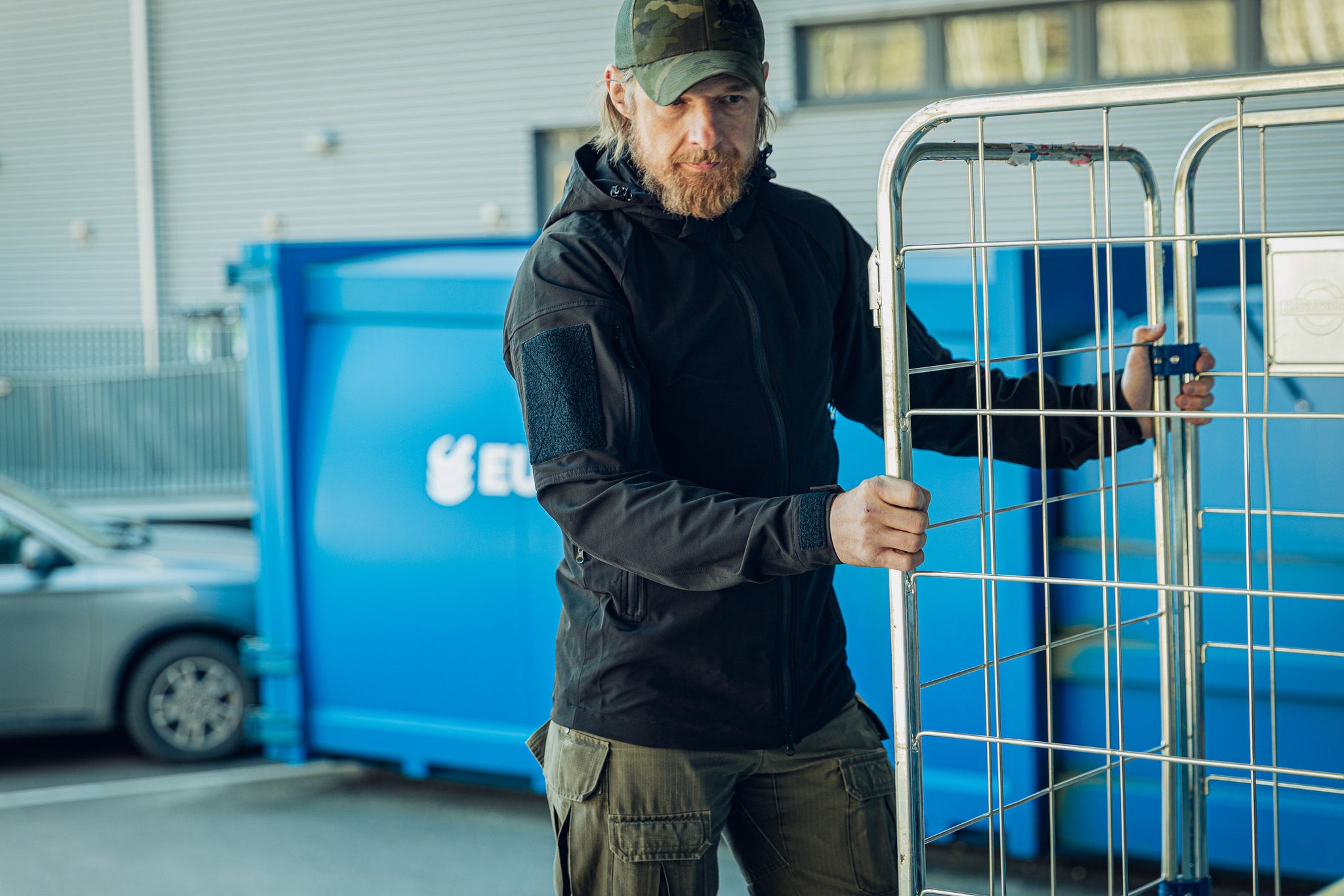 Man holding a metal cage in front of a blue recycling bin wearing a black jacket.
