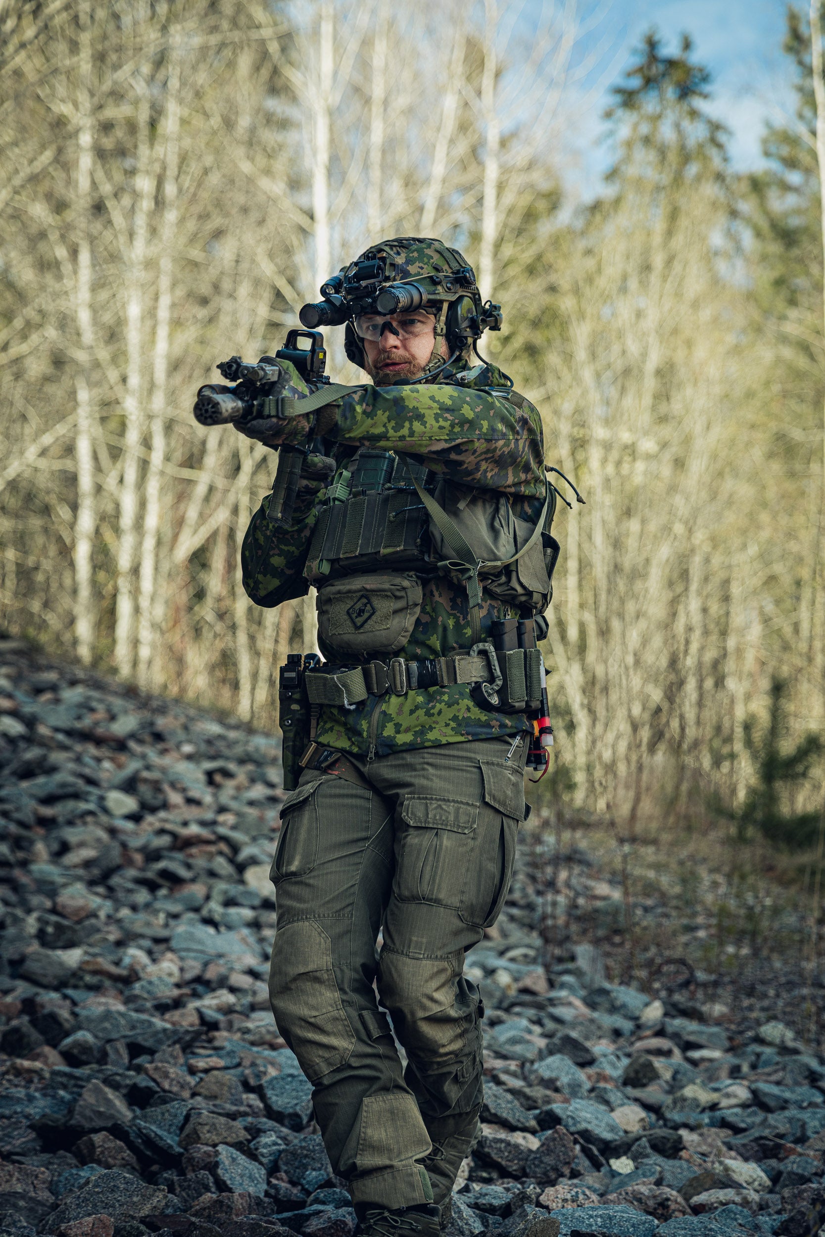 Person in camouflage military gear holding a rifle walking down hill on a rocky path with a forest in the background.