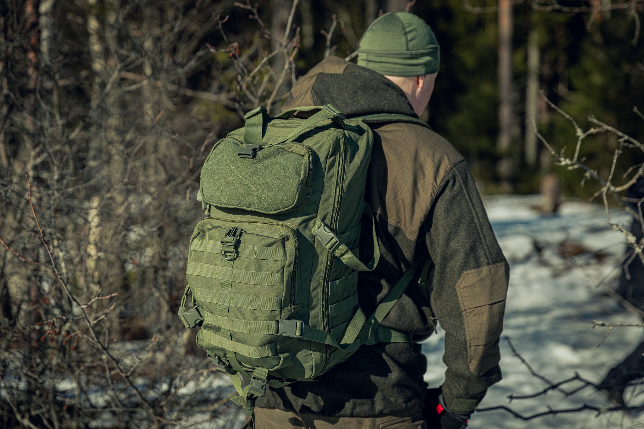 A backshot of a man in a snowy forest wearing green clothing and a green backpack. The sun is shining and he seems to be walking away from the camera.
