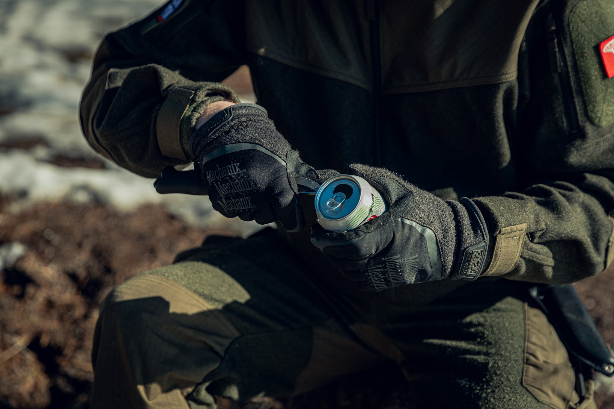 A close-up of a person holding a knife in one hand and a tin can in the other. The person appears to be slicing the top of the can open. They are wearing green clothing, and you can see some blurry snow and brown earth in the background.