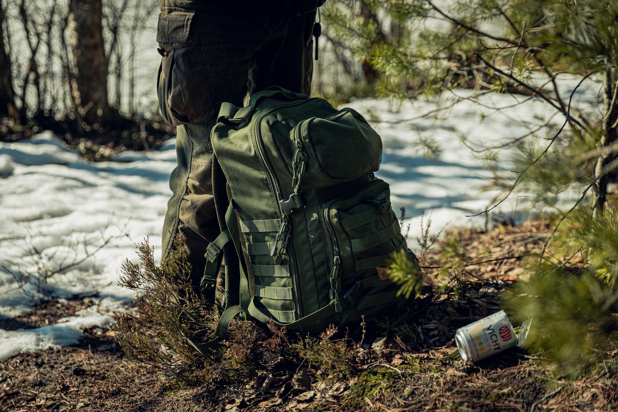 A green backpack on the ground in a forest with some snow in the background. The backpack is leaning against a person's legs. There is a cut-open can of sparkling water on the ground.