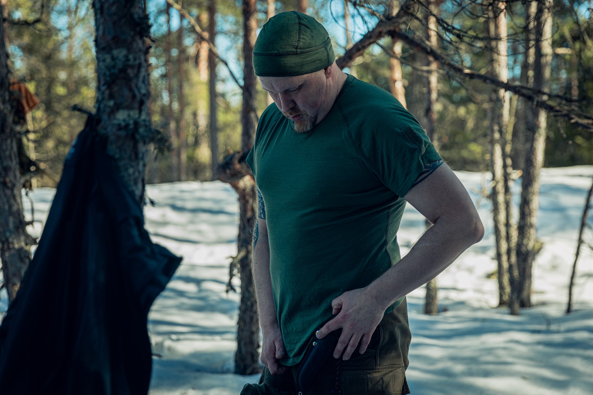 A man in a sunny and snowy pine wood forest, wearing green pants, a green t-shirt, and a green beanie. 