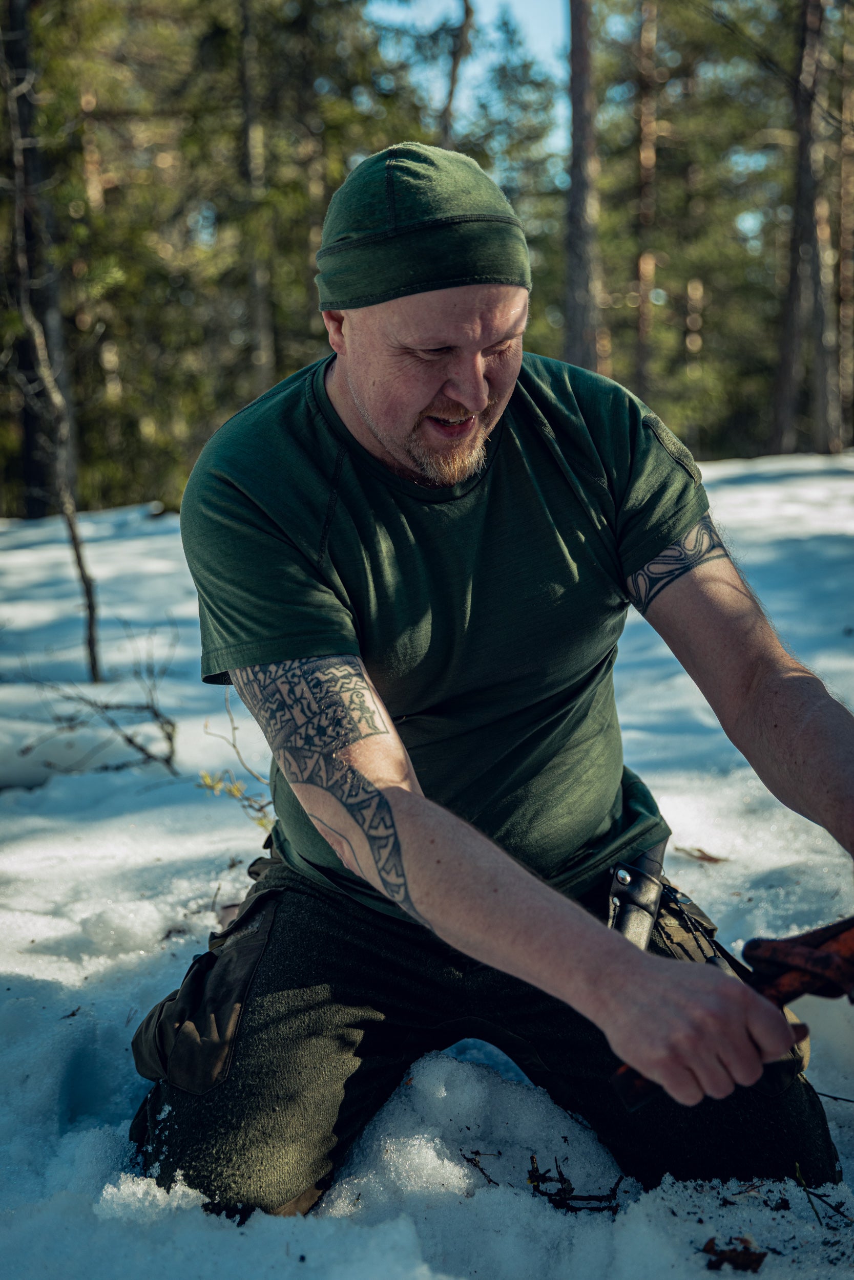 A man in a sunny and snowy pine wood forest, kneeling in the snow with an axe in his hands. He is wearing green pants, a green t-shirt, and a green beanie. He is holding some equipment in his hands, maybe the handle of a saw.