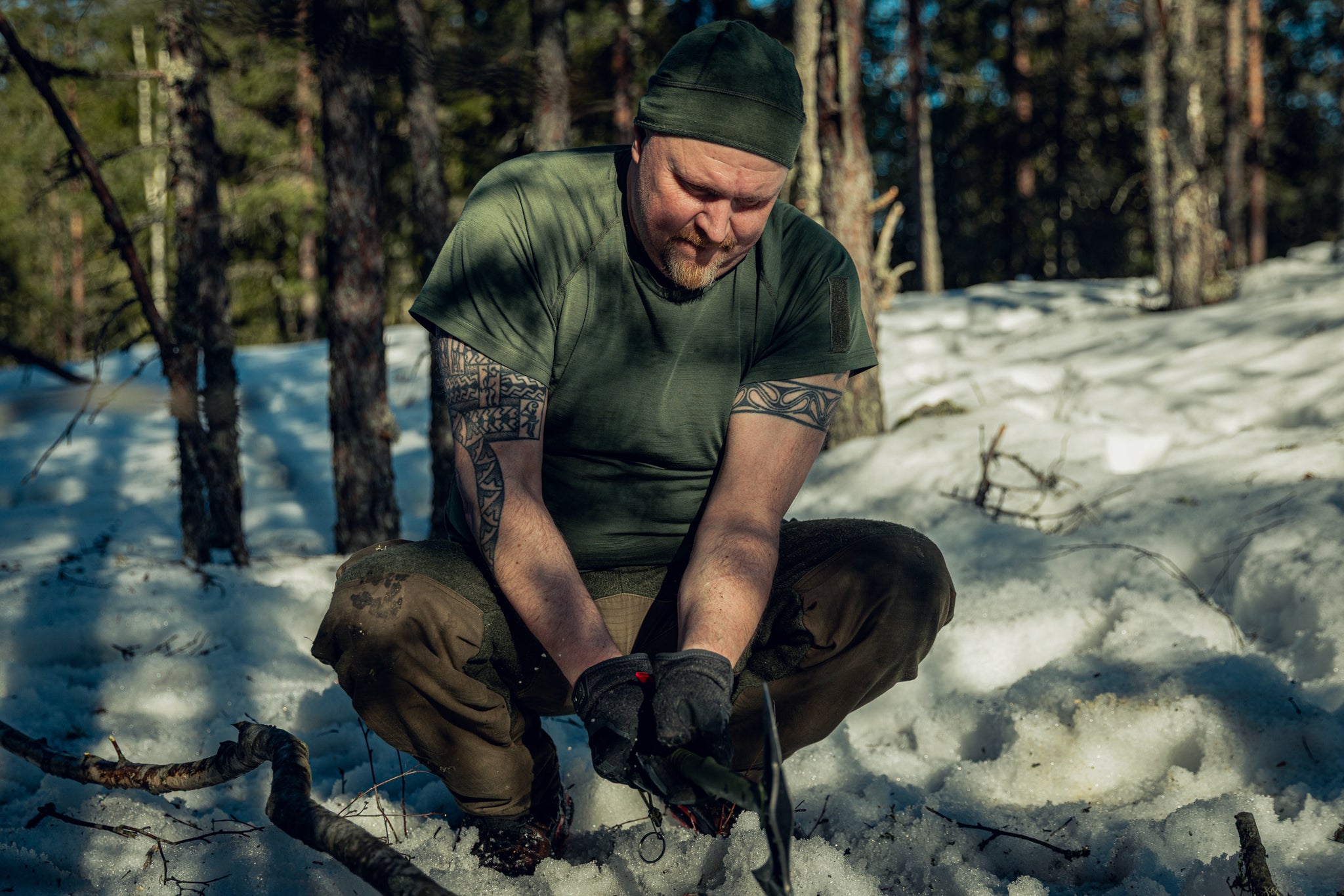 A man in a sunny and snowy pine wood forest, squatting in the snow with an axe in his hands. He is wearing green pants, a green t-shirt, black gloves and a green beanie.