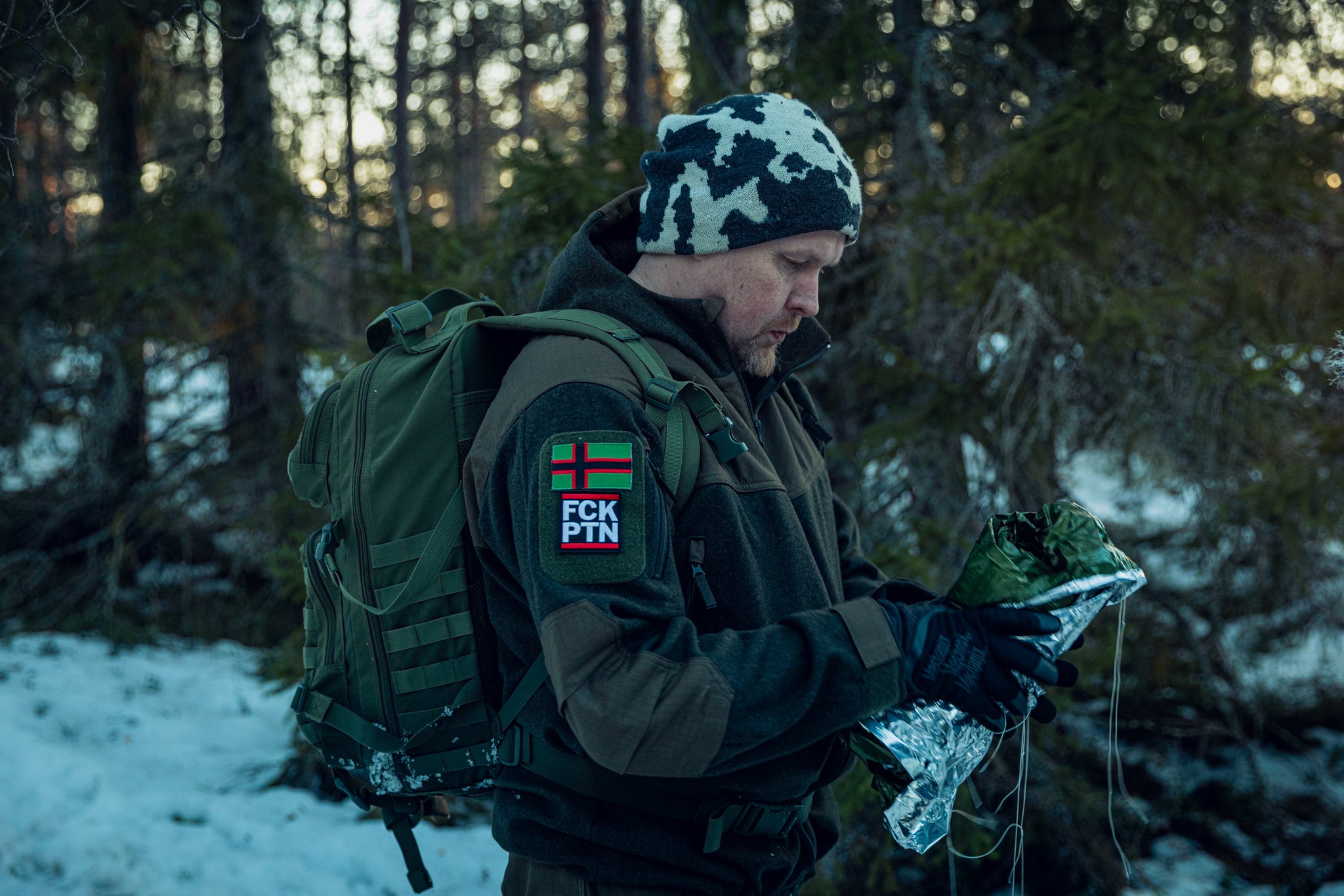 A man wearing a green jacket, snow camo cap and a green packpack in a snowy pine forest. He is holding a  folded-up space blanket in his hands, and he has a red-green-black Karelia flag patch and a patch that says FCK PTN on his jacket sleeve.