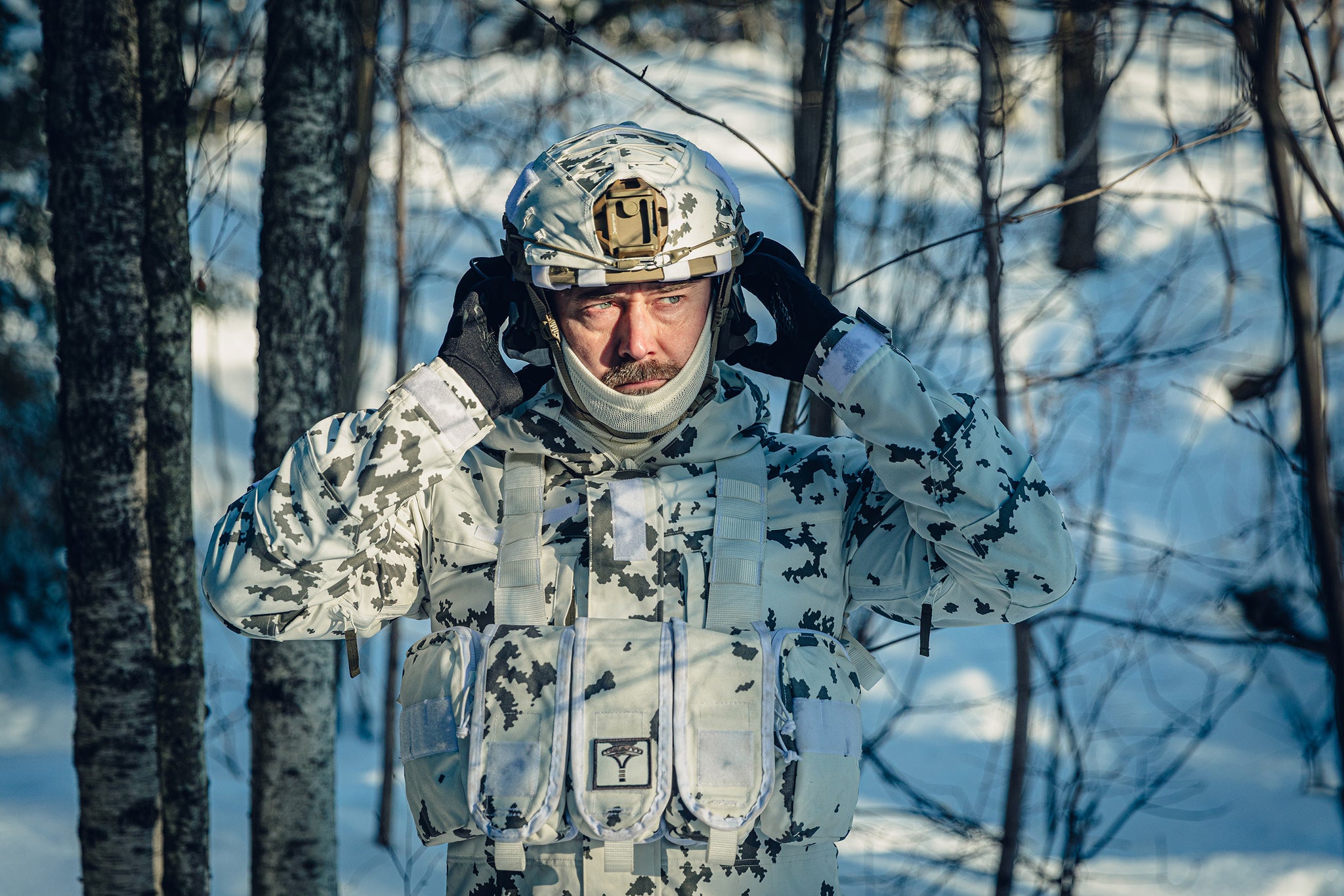 A man in a sunny, snowy forest wearing a Finnish M05 snow camo uniform and a combat helmet.  He is adjusting his hearing protection and there is a white neck tube covering part of his chin.