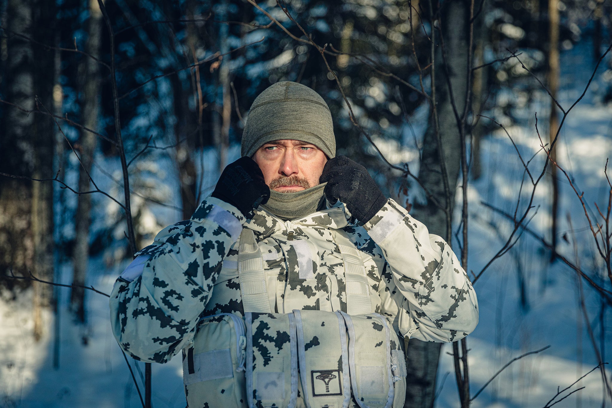A man in a sunny, snowy forest wearing a Finnish M05 snow camo uniform and a grey balaclava.  He is adjusting his balaclava and looking sternly to the left.