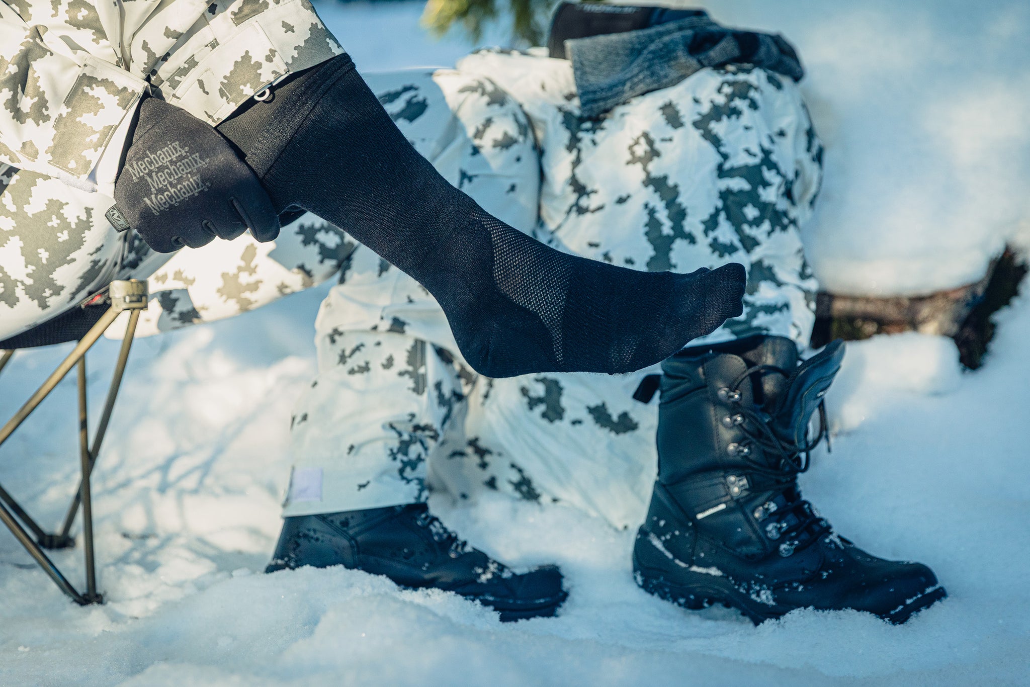 Close-up of a foot in a black sock with some mesh details. The person is sitting in the snow and wearing snow camo clothing, pulling at the sock with gloved hands.
