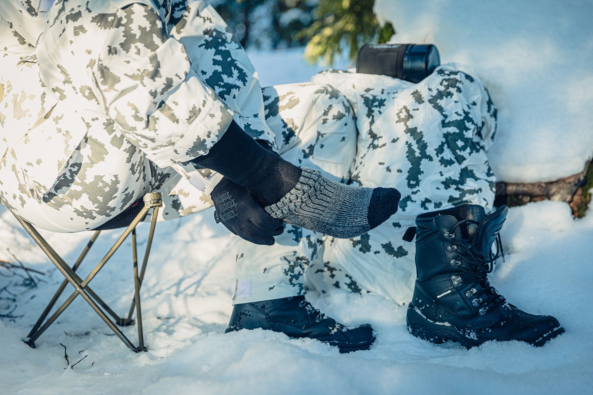 A person is sitting on a stool in the snow, pulling a grey sock on top of a black sock. The person is wearing snow camo clothing.
