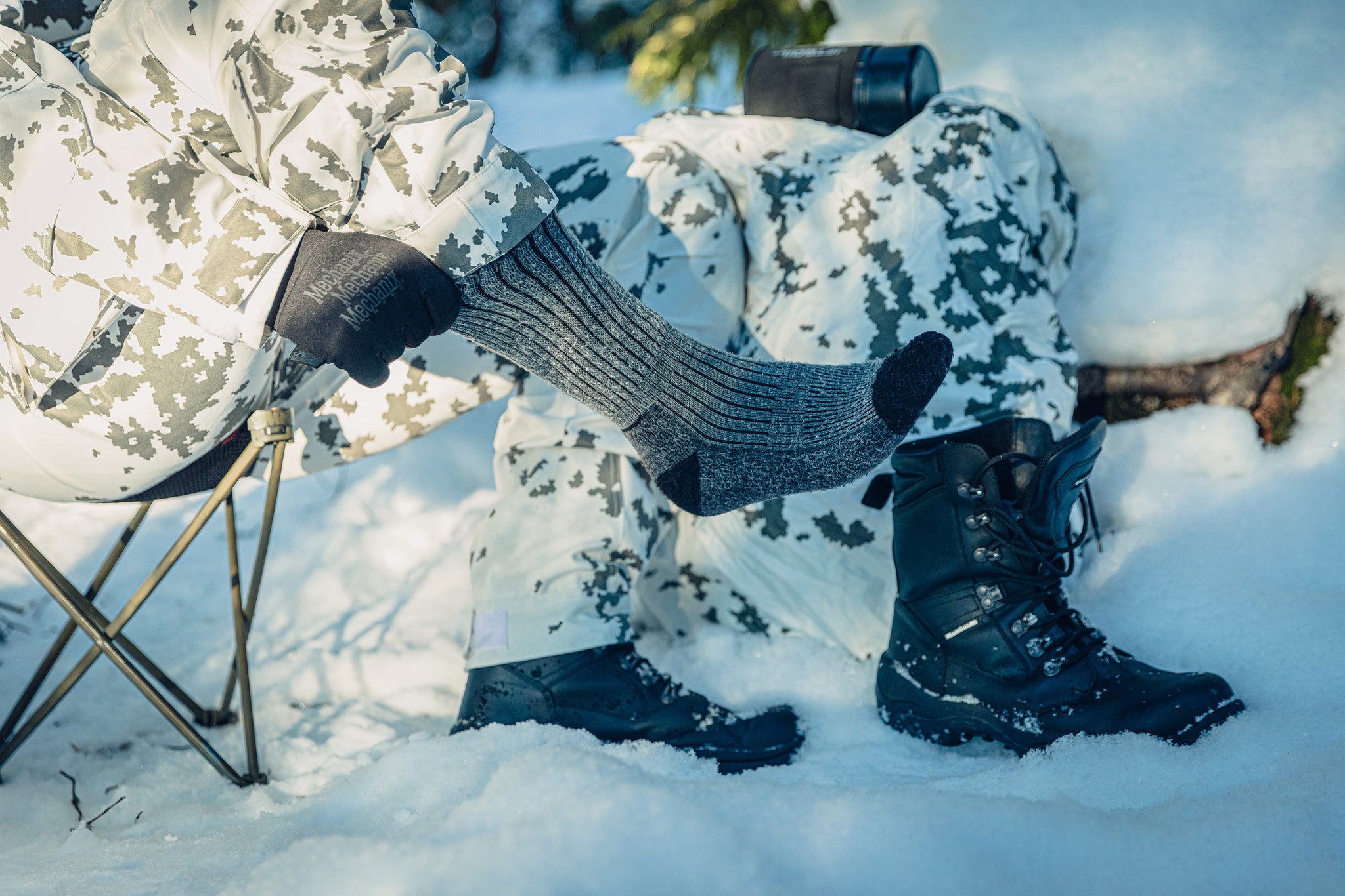 Close-up of a foot in a grey striped sock. The person is sitting on a stool in the snow and wearing snow camo clothing, pulling at the sock with gloved hands.
