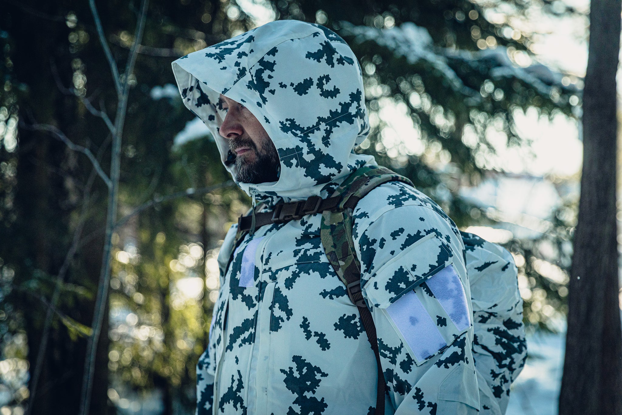 A man in a sunny, snowy forest wearing a Finnish M05 snow camo jacket with the hood up and a multicam backpack.  He is looking to the left.