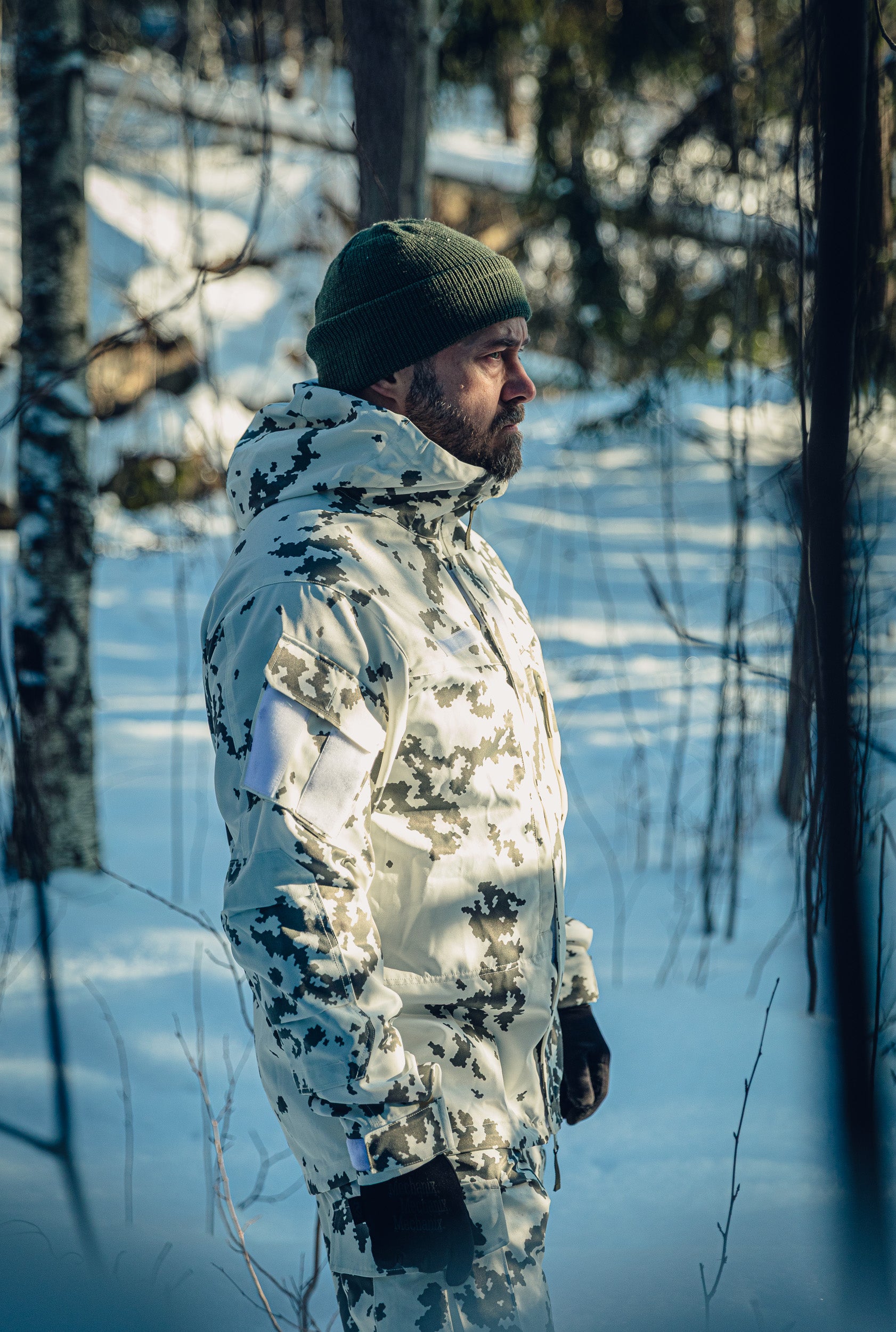 A man in a sunny, snowy forest wearing a Finnish M05 snow camo uniform jacket and pants, and green beanie. 