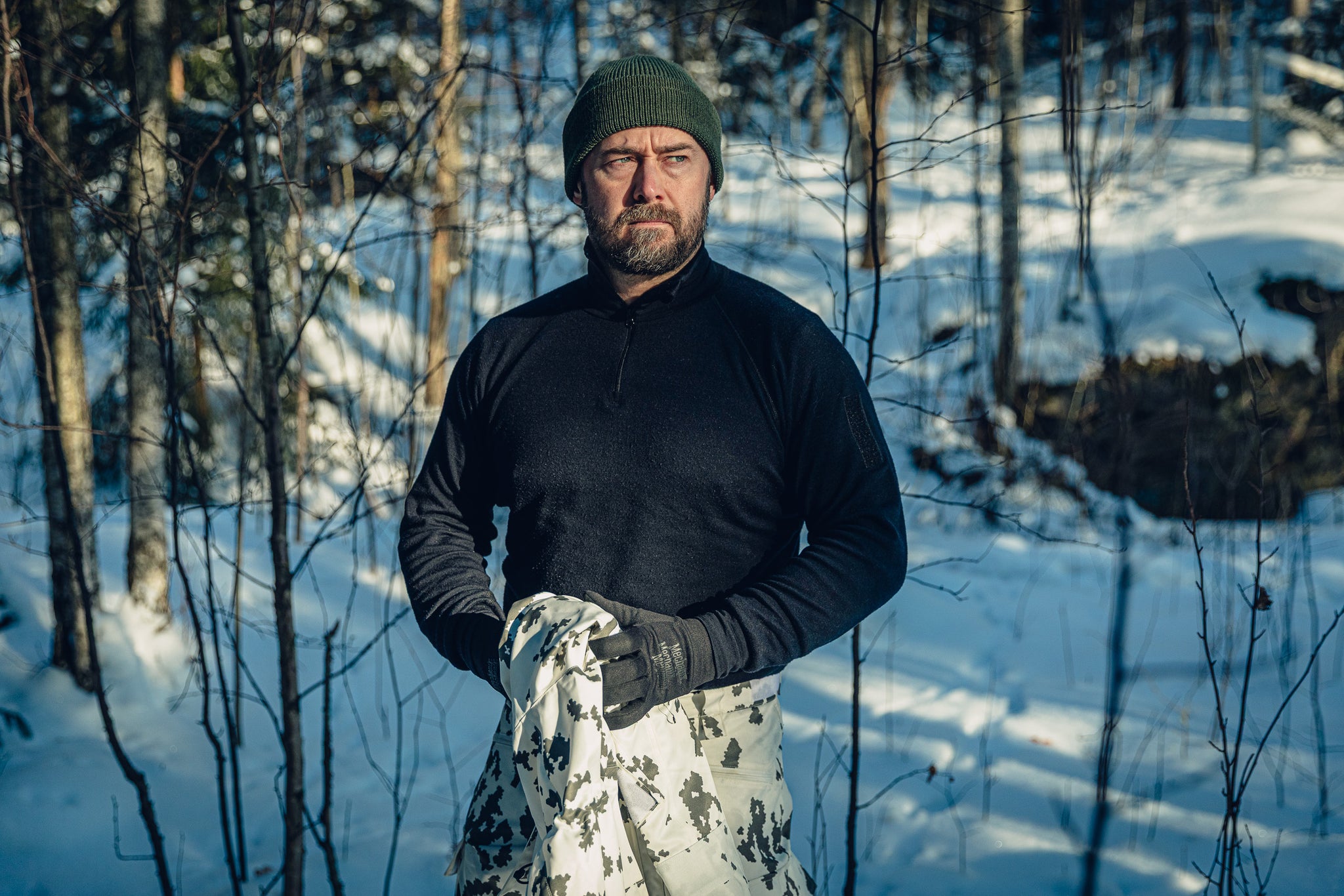 A man in a sunny, snowy forest wearing Finnish M05 snow camo pants, a black turtleneck shirt, and a green beanie. He is holding a snow camo garment in his hands and looking to the right.