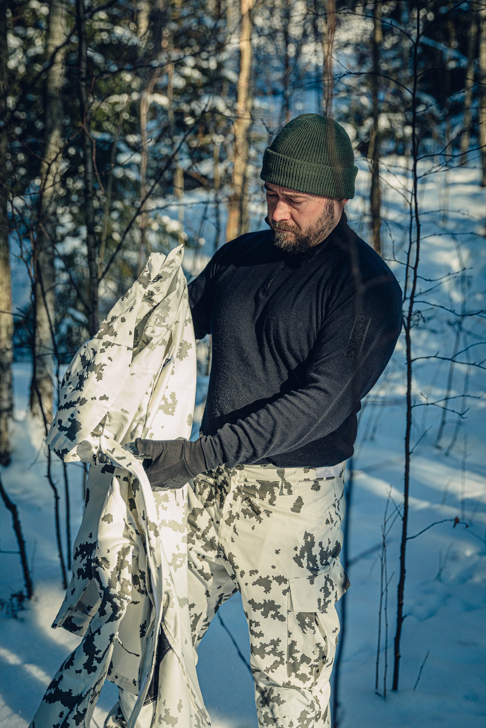 A man in a sunny, snowy forest wearing Finnish M05 snow camo pants, a black polo shirt and a green beanie. He is holding a snow camo jacket in his hands and looking down at it.