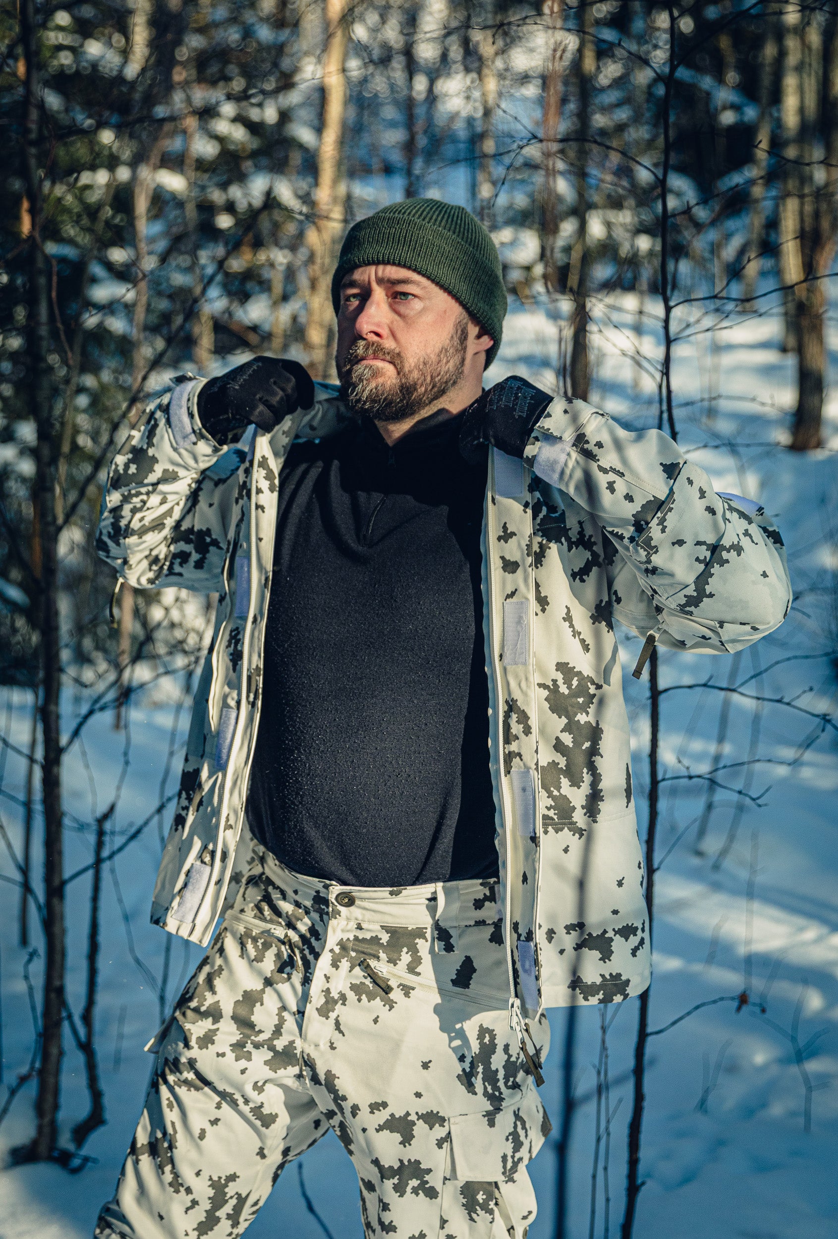A man in a sunny, snowy forest wearing a Finnish M05 snow camo jacket and pants, black turtleneck shirt and green beanie. The jacket is unzipper and he is holding the sides of it as if to adjust it.