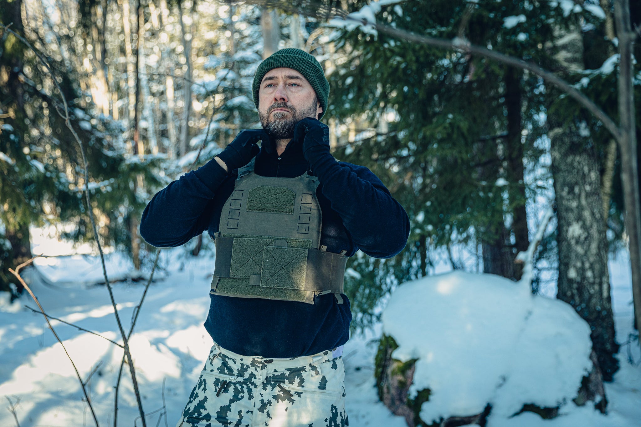 A man in a sunny, snowy forest wearing Finnish M05 snow camo pants, a black long-sleeved turtleneck shirt, a green beanie and a green plate carrier. He is adjusting the collar of his shirt.