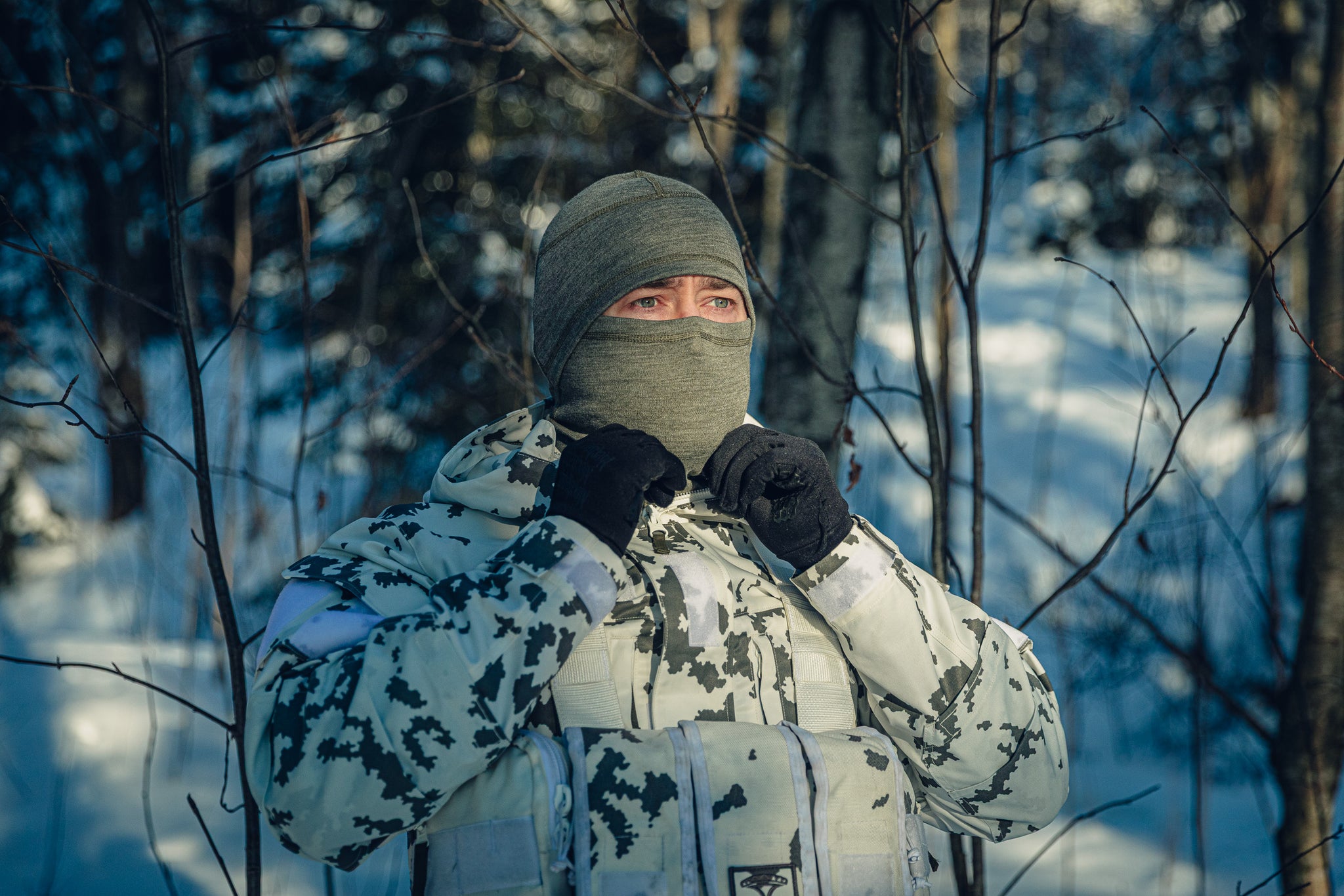 A man in a sunny, snowy forest wearing a Finnish M05 snow camo uniform and a grey balaclava.  He has pulled the balaclava to cover his entire face, only his eyes are visible.