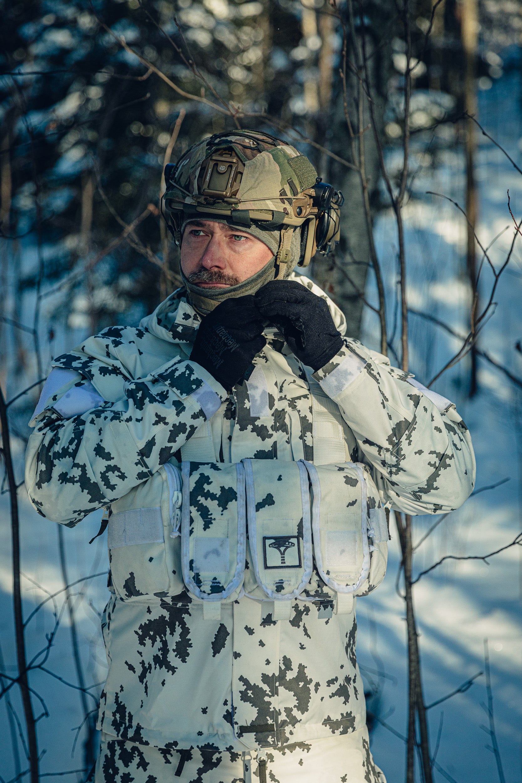 A man in a sunny, snowy forest wearing a Finnish M05 snow camo uniform and a combat helmet.  He is adjusting the strap of his helmet, and there is a grey balaclava under the helmet.