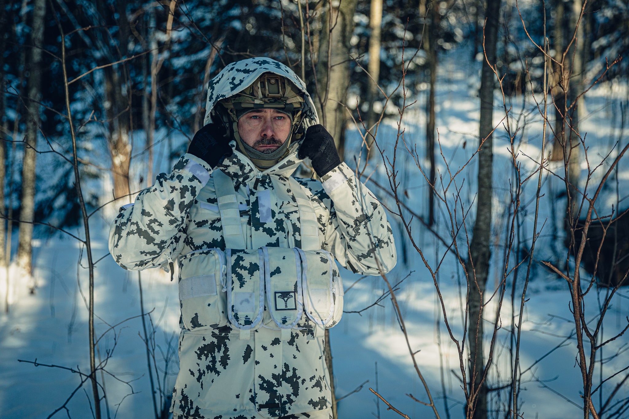A man in a sunny, snowy forest wearing a Finnish M05 snow camo uniform and a combat helmet.  He is adjusting the hood of the jacket, which is on top of the helmet, and there is a grey balaclava under the helmet.