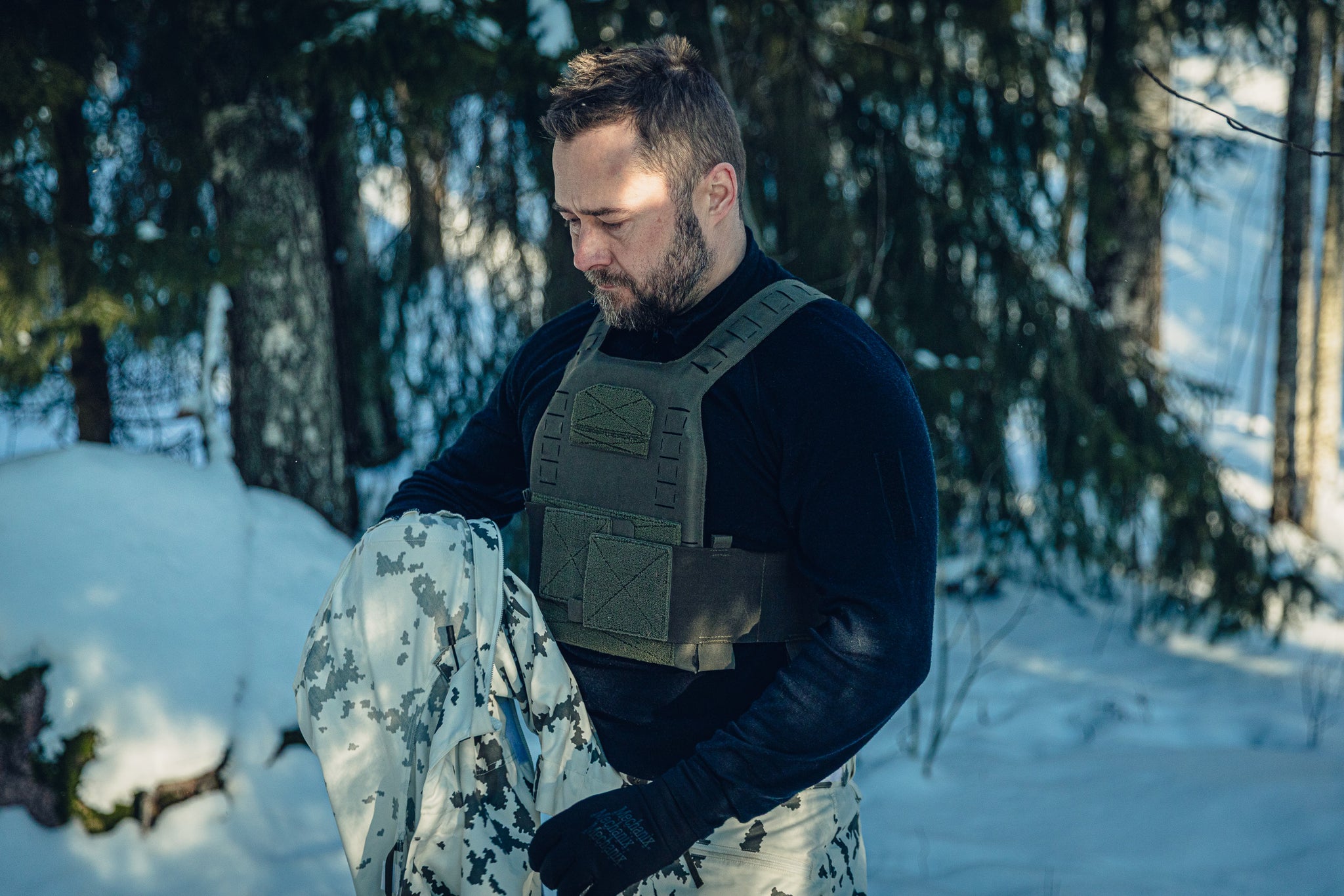 A man in a sunny, snowy forest wearing Finnish M05 snow camo pants, a black long-sleeved polo shirt, a green plate carrier. He is holding another snow camo garment in his hands and looking down.